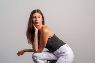 Moda mankeni stüdyodaki bir taburede zarif bir şekilde oturuyor, yumuşak gri bir arka plana karşı fotoğraf çekimi için poz veriyor, zarafet ve kendine güven yayıyor.