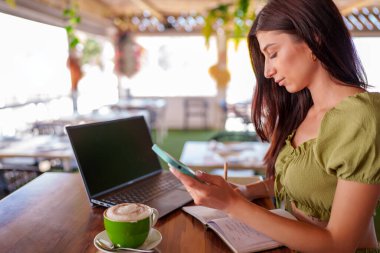 Akıllı telefonunu kullanan ve not defterine not alan genç bir kadın. Masasında dizüstü bilgisayar ve kapuçino var.