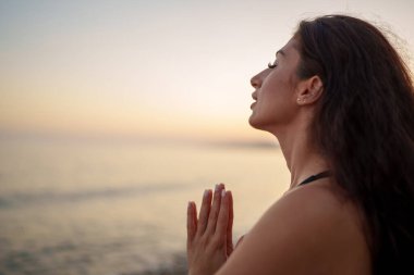 Meditasyon yapan bir kadın, elleri birbirine bastırılmış, altın gün batımı ışığı sırasında okyanusun yanında iç huzuru bulan bir kadın.