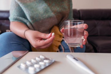 Kadın suyla hap alıyor, hastalıkla uğraşıyor. Masadaki termometre ve ilaçlar sağlık ve hastalık kavramlarını vurguluyor