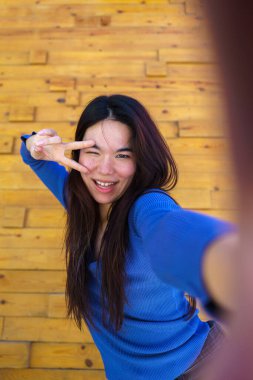 Neşeli genç Japon kadın zafer işaretini parmaklarıyla yaparken selfie çekiyor. Tahta bir arka planda göz kırpıyor ve gülümsüyor.