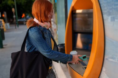 Kızıl saçlı kadın otomatik veznedar servisinden cep telefonuyla faydalanıyor ve dışarıda bir çanta taşıyor.