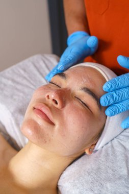 Kanepede uzanmış, yüz bakımı gören genç bir kadın. Bir güzellik uzmanının eldivenli elleri yüzüne ürün uyguluyor.