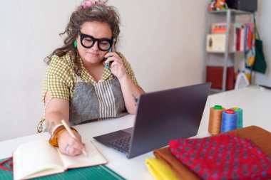 Odaklanmış kadın girişimci çevrimiçi iş yönetiyor, müşteriyle iletişim kuruyor ve evde dikiş dikerken notlar alıyor.