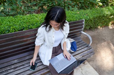 Genç bir kadın parkta bankta oturuyor, dizüstü bilgisayar kullanıyor, akıllı telefon tutuyor, dışarıda çalışmaktan zevk alıyor.