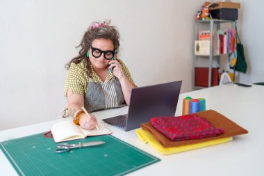 Terzi telefonla emir alıyor ve bir deftere yazı yazıyor. Parlak atölyesindeki küçük işletmesini yönetiyor.