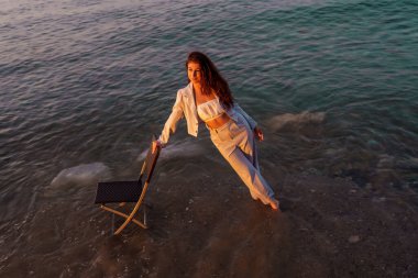 Çıplak ayaklı profesyonel bir kadın, deniz kenarındaki sandalyede dinleniyor, zarif iş kıyafetleriyle altın gün batımını seyrediyor.