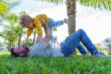 Baba çimenlerde yatıyor, oğluyla oynuyor. Onu kaldırıyor, parkta birlikte eğleniyor.