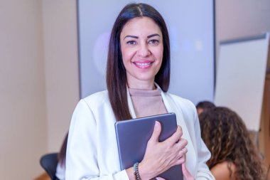 İş kadını gülümseyerek ve elinde tablet tutarak modern bir kongre merkezinde iş toplantısına katılıyor, güven ve profesyonellik yayıyor.