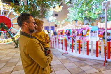 Oğlunu kucağında tutan baba, Noel pazarında atlı karıncayı seyrediyor, neşeli atmosferin tadını çıkarıyor.