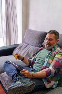 Sakallı, orta yaşlı bir adam evinde koltukta rahatça uzanmış tablet kullanırken gülümsüyor ve elinde sarı bir kredi kartıyla online alışveriş, bankacılık ve dijital yaşam tarzını temsil ediyor..