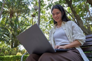 Gülümseyen genç Japon kadın parktaki bankta telgraf çekerken dizüstü bilgisayar kullanıyor, iş hayatının tadını çıkarıyor.