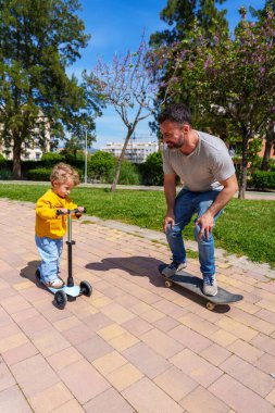 Baba kaykayla oğluyla birlikte parkta kayıyor, neşeli bir bağ kurma anını paylaşıyor.