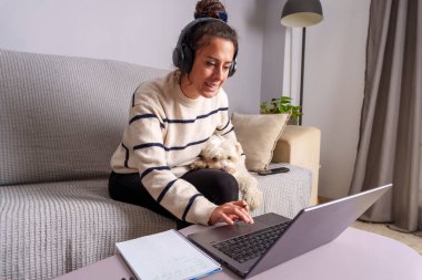 Kulaklık takan genç bir kadın evdeki kanepede yanında oturan köpeğiyle çevrimiçi kursa giderken dizüstü bilgisayar kullanıyor.
