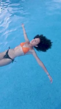 Parlak mavi yüzme havuzunda huzur içinde yüzen genç kadın, kollarını açıp yaz tatilinin tadını çıkarıyor, saf rahatlık ve özgürlüğün vücut bulmuş hali.