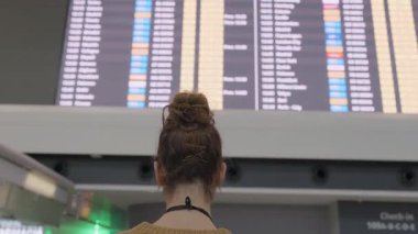 Kadın gezgin arkasını döndü, modern havaalanı terminalindeki dijital zaman çizelgesini tarıyor, uçuş bilgilerini ve tarifesini kontrol ediyor.