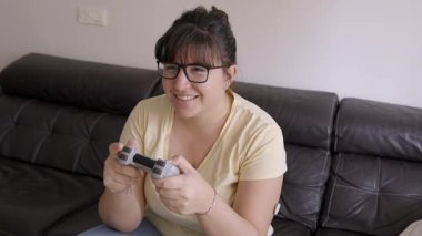 Hevesli genç bir kadın dikkatle oyun oynuyor, kumandayı tutuyor ve rahatça oturma odasındaki koltukta otururken zaferi kutluyor.
