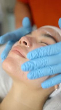 Güzellik uzmanı nemlendirici krem sürüyor, mavi eldiven giyiyor, beyaz havluyla dinlenen genç Japon kadının cildine masaj yapıyor.