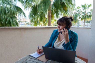 Uzaktan çalışan profesyonel bir kadın, açık hava balkonunda verimli tropik yeşillikler arasında notlar yazarken cep telefonuyla konuşuyor.