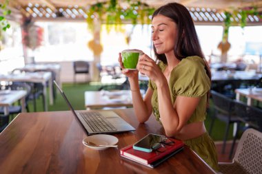 Sıcak bir restoran masasında laptopuyla çalışırken kapuçino içen genç bir kadın yaz sabahının sükunetini kucaklıyor.