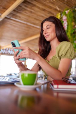 Bir kafede uzaktan çalışırken akıllı telefon ve dizüstü bilgisayar kullanan genç bir kadın bir fincan kahvenin tadını çıkarıyor.