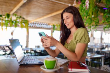 Profesyonel yönetici ağı, yeşil dekorlu modaya uygun bir kafede dizüstü bilgisayar ve akıllı telefon üzerinde çalışırken cappuccino içmek