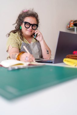 Terzi telefonda konuşurken not alıyor ve dikiş atölyesinde dizüstü bilgisayar kullanıyor, siparişleri ve müşteri hizmetlerini yönetiyor.