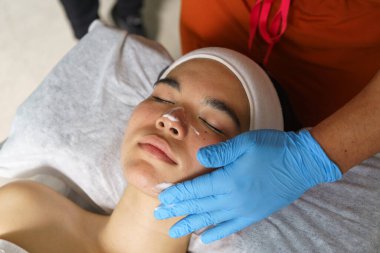 Mavi eldiven giyen bir güzellik uzmanı beyaz havluya uzanmış genç bir kadının yüzüne temizleyici köpük sürüp ultrasonik yüz temizleme ameliyatına hazırlanıyor.