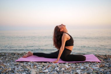 Çakıl taşı plajında yoga yapan kadın, altın gün batımı ışığı, yavaşça okyanus dalgaları arasında sakin meditasyonu yansıtıyor.