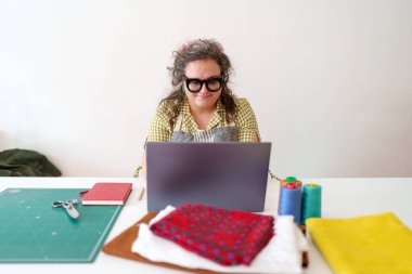 Dizüstü bilgisayarda yazan profesyonel terzi, canlı tekstil, dikiş iplikleri, kesme araçları, moda tasarım siparişleri ile çevrili.