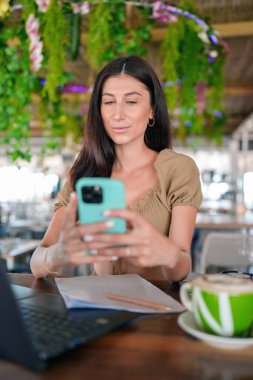 Akıllı telefon tutan genç bir kadın, yeşil dekore edilmiş bir kafede ekrana bakıyor, masada dizüstü bilgisayar ve kahve fincanı var.