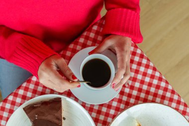 Kırmızı kazaklı bir kadın çay kaşığıyla bir fincan koyu kahveyi karıştırıyor ve kareli masa örtüsünde bir dilim çikolatalı kek yiyor.