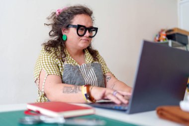 Uzaktan çalışan profesyonel bir kadın, ofis ortamında önlük ve gözlük takarken dizüstü bilgisayarda daktilo kullanıyor.