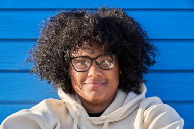 Güneşli bir günde mavi arka planda bereli kazak ve afro saç giyen siyahi bir kadın. 