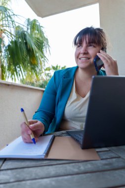 Kadın profesyonel ev terasından uzaktan çalışıyor, akıllı telefon ve defterle birden fazla iş yapıyor, neşeli davranışlar sergiliyor.