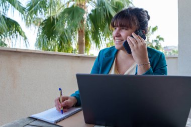 Profesyonel kadın çoklu görev, dizüstü bilgisayarla güneşli bir terastan cep telefonuna yazma ve arama