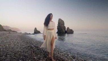 Çakıl taşlı kıyı şeridi boyunca çıplak ayakla yürüyen zarif bir model, sakin bir akşam ışığını saran sakin bir ifadeyle ve akan beyaz elbiseyle