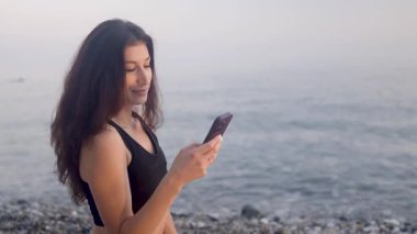 Genç bayan, sakin bir yaz akşamı sahile inerken akıllı telefondan keyif alıyor, doğal kıyı manzarasının ortasındaki dijital bağlantıyı yakalıyor.
