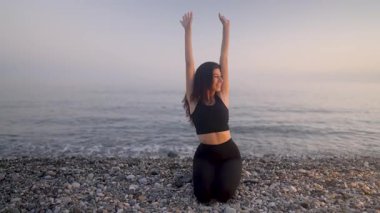 Huzurlu bir kadın, altın gün batımı ışığında kumlu deniz kıyısında yoga yapıyor. Sağlıklı ve dingin bir zindelik ve farkındalığın vücut bulmuş hali.