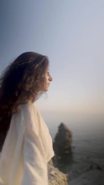 Huzurlu kadın silueti, ılık şafak ışınlarını emer, kayalık kıyı çıkıntısında çıplak ayakla durur, huzur verici bir güç ve kişisel özgürlük yayar.