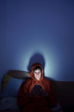 Gece yatağında cep telefonu kullanan bir genç. Ekran ışığıyla aydınlatılmış.