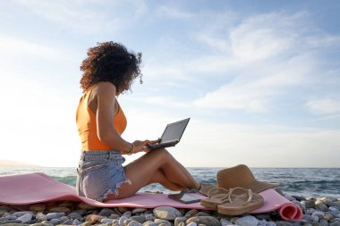 Bir çakıl taşı plajında dizüstü bilgisayarda çalışan genç bir kadın, doğanın ve okyanus kenarında esnek bir yaşam tarzının tadını çıkarıyor.