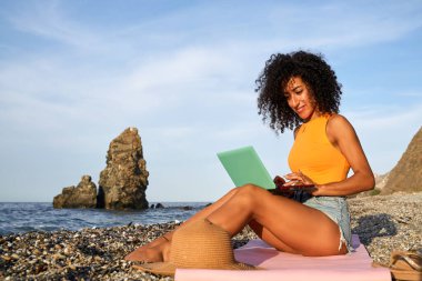 Güzel deniz ve kaya oluşumlarıyla çevrili çakıl taşı plajındaki bir paspasın üzerinde otururken dizüstü bilgisayar kullanan kıvırcık saçlı bir kadın.