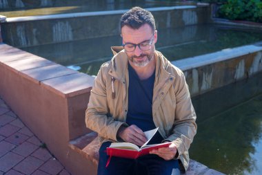 Gözlüklü bir adam kırmızı kitap ya da günlük okuyor, sakin bir su özelliğinin yanında oturuyor, düşüncelere dalmış ve öğrenmeye dalmış.