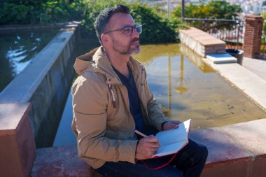 Olgun bir adam gözlük takıyor, su kenarında otururken not alıyor açık havada beyin fırtınası yapıyor ve yansıtıyor.