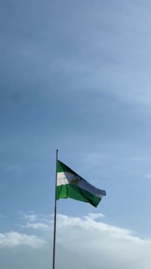 Endülüs özerk toplumunun resmi bayrağı bayrak direğinde sallanıyor. Arkasında bulutlar olan açık mavi bir gökyüzü.