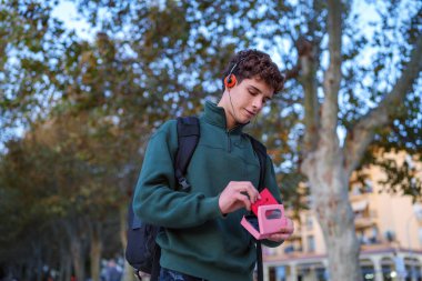 Genç adam dışarıda duruyor, turuncu kulaklıklar ve kasetle klasik bir müzik setinde müzik dinliyor.