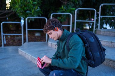 Kulaklık ve sırt çantası takan, basamaklarda oturan, müzik dinlemek için müzik aletini hazırlayan genç bir adam.