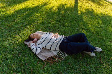 Kulaklıkla çimenlerde uzanan, müzik dinleyen, doğada huzur bulan, güneşlenen ve dinlenen genç bir kadın.