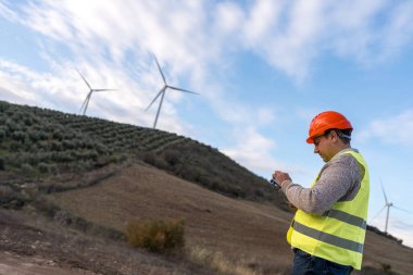 Uzaktan kumandalı bir uzaktan kumandalı çelik yelek ve başlık takan mühendis türbinli bir rüzgar çiftliğini inceliyor.